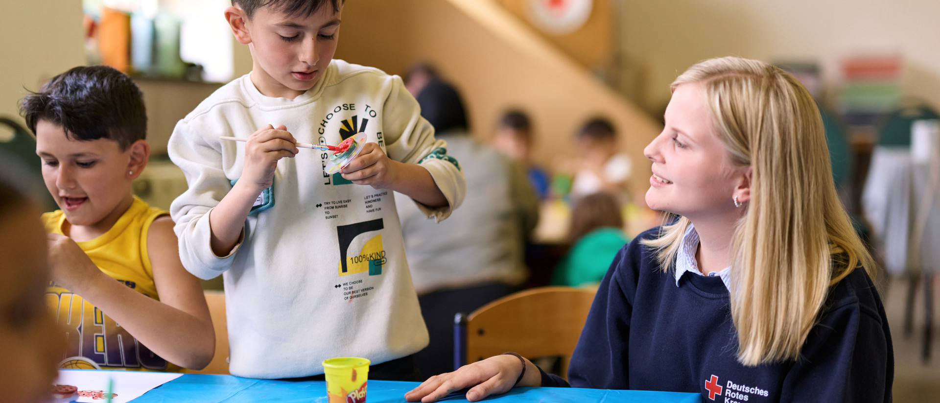 Kinder DRK-Freiwillige begleitet Kinder beim gemeinsamen Malen und Basteln an einem Tisch.